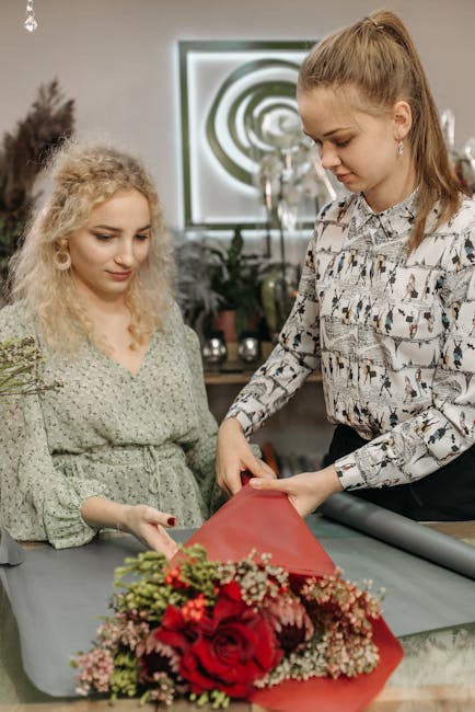 A florist with long, light brown hair tied in a high ponytail is carefully wrapping a floral bouquet, featuring vibrant red roses, pink and white gypsophila, and green foliage, in red wrapping paper.