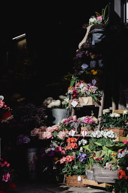 A vibrant display of fresh flowers arranged in various sizes of baskets and buckets, featuring pink, white, and purple blooms including roses, peonies, and lisianthus. The flowers are set outdoors wit