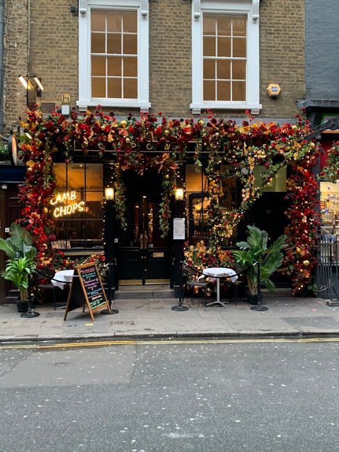 Exterior view of a cozy pub or restaurant on West End Lane in West Hampstead, decorated with an abundant floral display of red, pink, and yellow flowers climbing around the black entrance door and abo